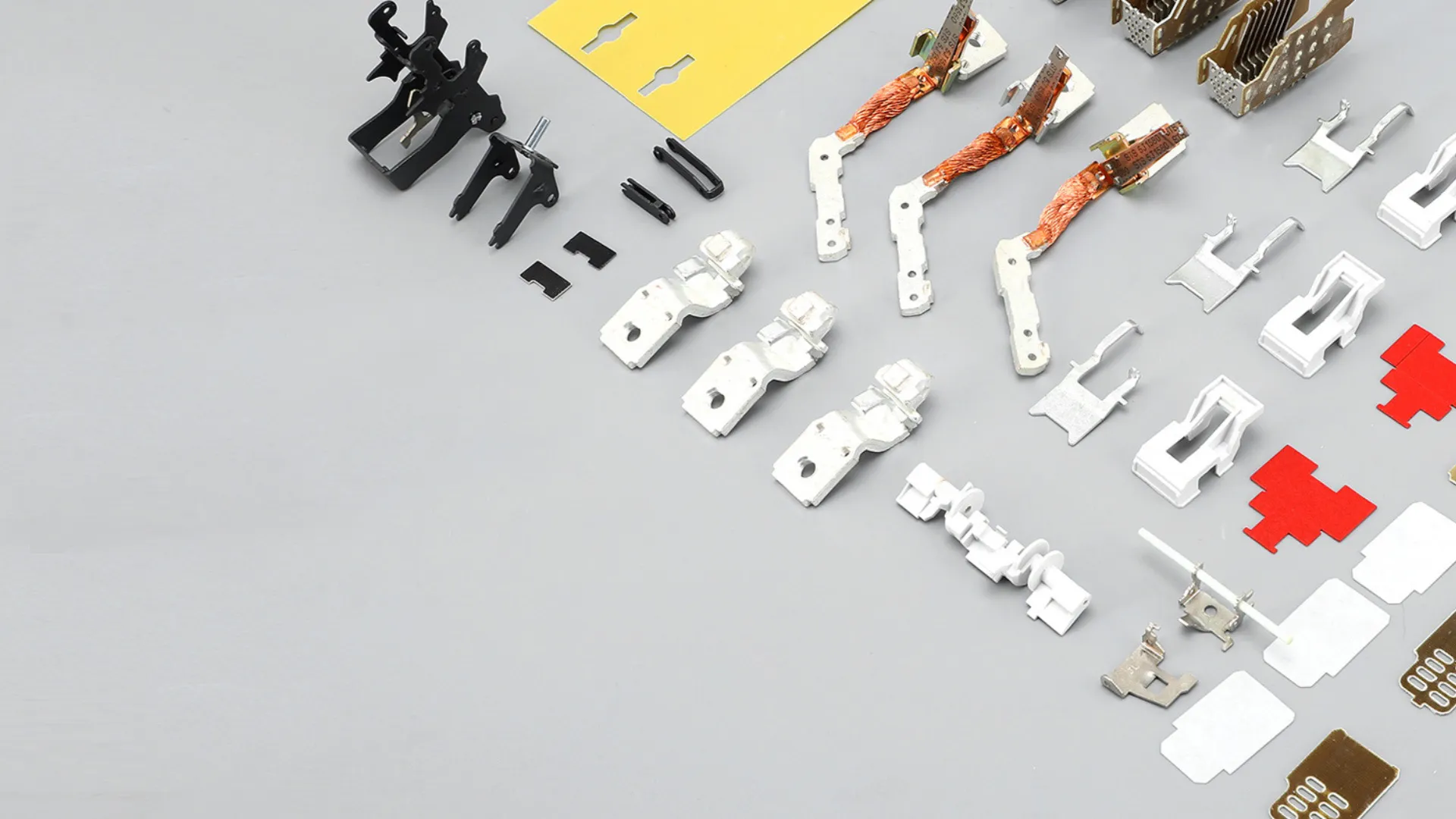 Various internal parts of a molded case circuit breaker laid out on a light grey surface, including black plastic levers, white plastic mechanisms, copper coil assemblies, metal terminals, and red and yellow plastic pieces.