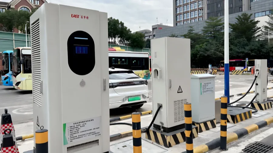 White electric vehicle charging cabinets with cables and bollards in a parking area, with cars and buses in the background.