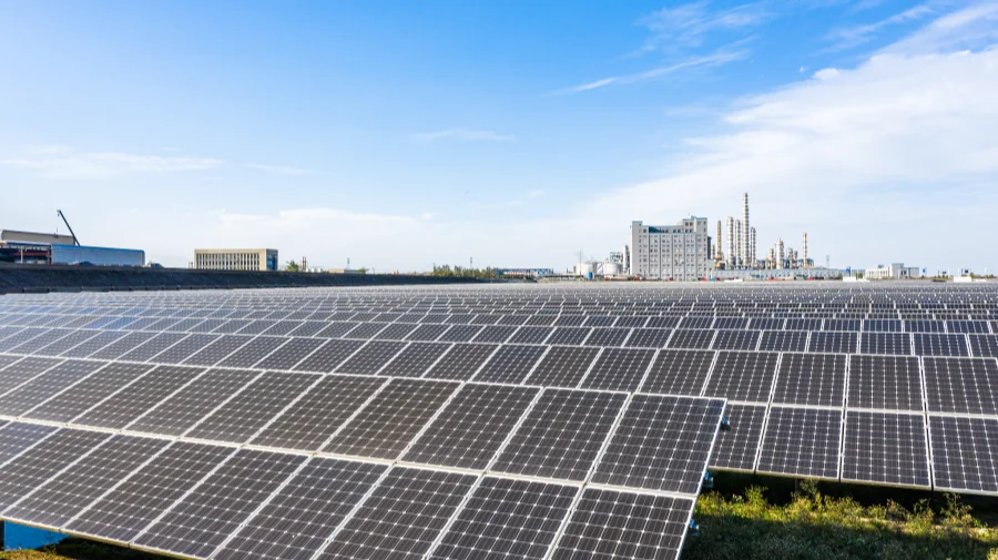 A large field of solar panels under a blue sky, with industrial factory buildings visible in the distance.