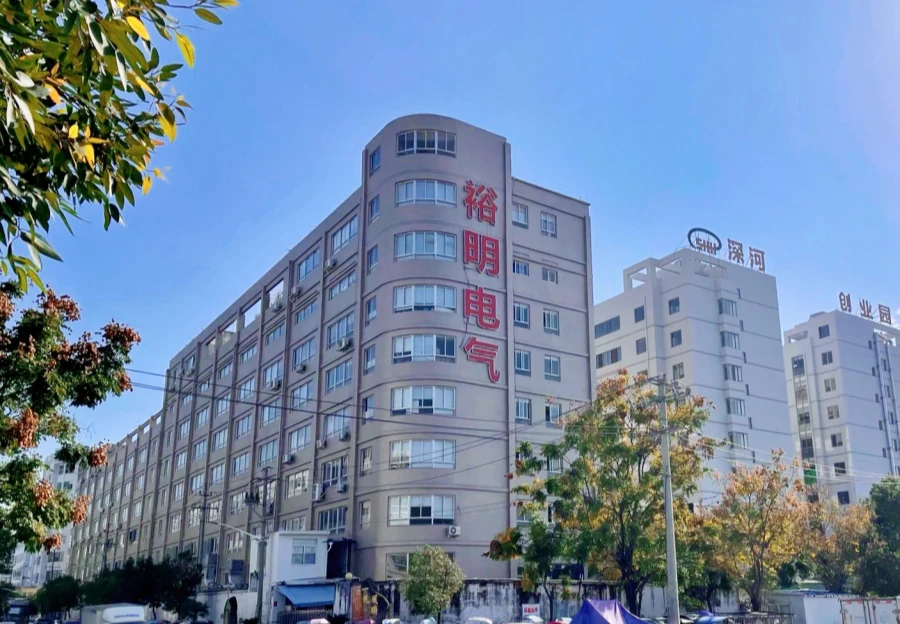 A multi-story beige factory building for Yumonn Electric, with red Chinese characters on its curved corner, next to a white building under a clear blue sky.